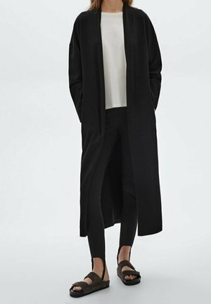 Personne portant un long manteau noir ouvert, un haut blanc, un pantalon noir ajusté avec des étriers et des sandales sombres slip-on, debout devant un fond uni.
