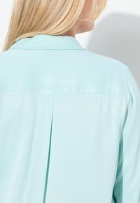 Mintgrøn bluse med en glat tekstur, klassisk krave og et plisseret design bagpå for ekstra detaljer. Stoffet falder blødt.