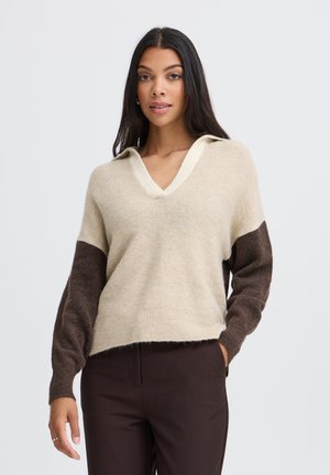 Femme aux longs cheveux foncés porte un pull beige à col en V avec des manches marron et un pantalon marron foncé, debout sur un fond clair uni.