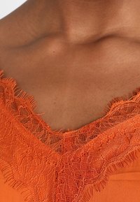 Orange blus med en djup V-ringning prydd med intrikata spetsdetaljer. Tyget verkar slätt med en mjuk textur.