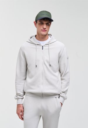 Ung mand iført en lysegrå hættetrøje med lynlås, matchende joggingbukser, hvid T-shirt og en grøn baseballkasket, stående foran en ensfarvet baggrund.