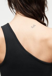 Górna część pleców kobiety z ciemnymi włosami opadającymi na ramię, ubrana w czarny, prążkowany top na jedno ramię, przy prawym łopatce widoczny mały tatuaż kota.