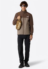 Giacca in pile con cerniera marrone e beige a maniche lunghe, abbinata a pantaloni neri e stivali scuri. Una piccola borsa a tracolla beige chiaro è indossata a tracolla.