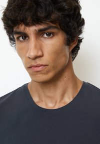 Donkerblauw rondenek t-shirt op een persoon met kort, krullend haar. Nabij opname van gelaatstrekken, inclusief een zachte huid en een neutrale uitdrukking.