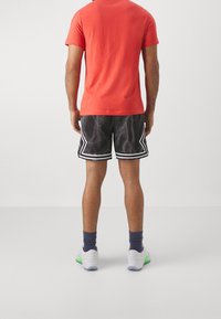 Rotes T-Shirt mit kurzen Ärmeln kombiniert mit schwarzen Shorts, die ein Blitzmuster und weiße Akzente aufweisen, sowie sportlichen Schuhen mit grünen Highlights.