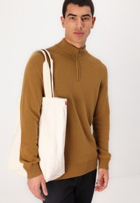 Maglione marrone con zip e collo a imbuto, con orlo e polsini a coste, abbinato a una borsa in tela color crema. La borsa presenta un'etichetta di accento rossa.