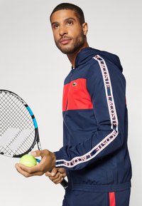 Marineblå og rød lynlåsjakke med hætte, med Lacoste-logo på brystet og ermerne. Modellen holder en tennisracket og bold.