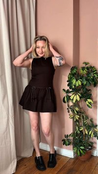 Νεαρή γυναίκα με μαύρο αμάνικο φόρεμα και μαύρα παπούτσια ποζάρει με τα χέρια στα μαλλιά δίπλα σε μια πράσινη γλάστρα με φυτό, μπροστά σε ροζ τοίχο.