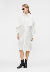 Žena oblečená v bielych priehľadných midi šatách na gombíky so širokými rukávmi, čiernych členkových čižmách a malých čiernych okuliaroch, stojaca pred bielym pozadím.