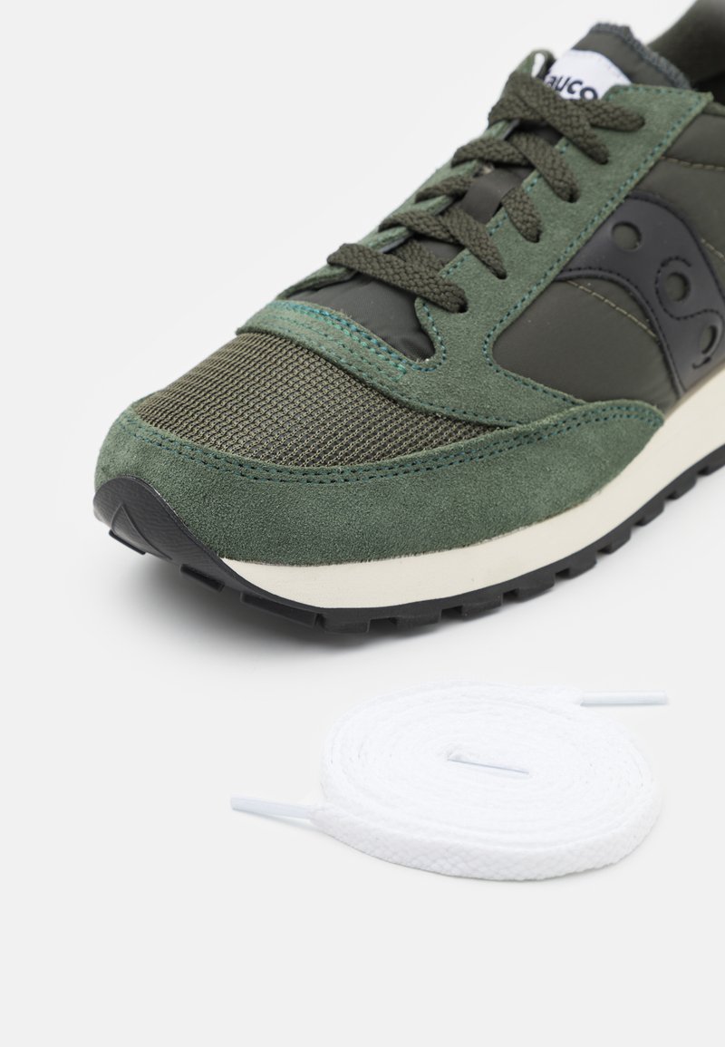 Zapatilla de deporte verde de ante y malla con acentos en negro, superposición texturizada y un cordón redondo blanco al lado. Suela de goma blanca con tracción.