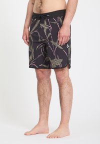 Homme portant un short de bain noir avec des motifs de feuilles vertes et roses, debout pieds nus devant un fond blanc uni.