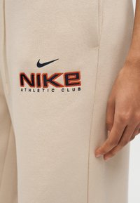 Μπεζ αθλητικό παντελόνι Nike με μαύρο και πορτοκαλί λογότυπο «Nike Athletic Club» κοντά στην τσέπη, και το χέρι ενός ατόμου τοποθετημένο δίπλα στο πόδι.