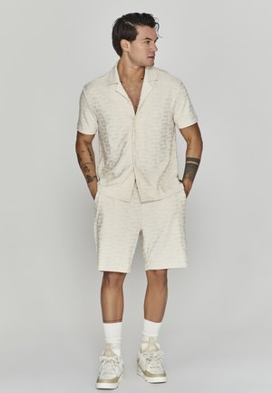 Camicetta crema a maniche corte e pantaloni corti coordinati con un sottile motivo geometrico; indossati con sneakers bianche e calze alla caviglia.