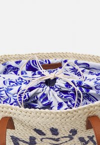 Kudottu olkilaukku, jossa on ruskeat nahkahihnat ja sinivalkoinen kukkakuvioinen sisävuori, joka on sidottu nauhalla.