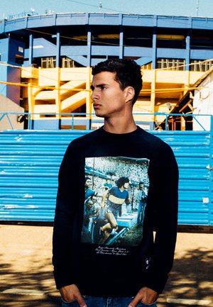 Zwarte lange mouwen shirt met een voorkantafbeelding van voetballers, met een blauw en gele stadionachtergrond. Glad materiaal, reguliere pasvorm.