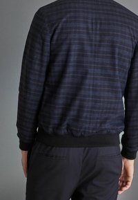 Navy blue jacket with a grid pattern, textured fabric, elastic cuffs, and waistband. Worn over dark pants with a side pocket detail.