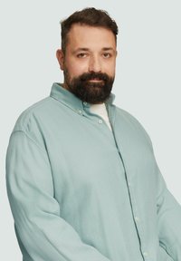 Lichtblauw gestructureerd overhemd met kraag, met een witte T-shirt zichtbaar eronder, gedragen door een man met een baard.