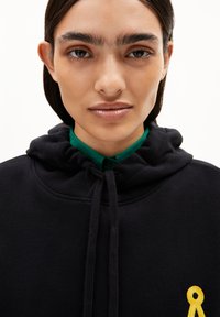 Felpa nera con cappuccio, decorata con ricami a nastro giallo. Il modello indossa una camicia verde con colletto sotto. Tessuto dalla superficie liscia.