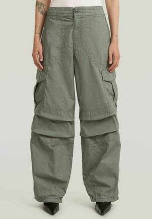 Pantalon cargo vert avec jambes amovibles zippées, grandes poches latérales, taille élastiquée, porté avec des chaussures noires pointues et des tatouages visibles sur les bras.