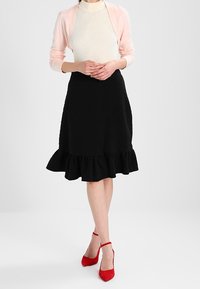 Falda negra texturizada con un dobladillo con volantes, combinada con una blusa blanca de canalé y un cárdigan rosa claro. Zapatos de tacón rojo completan el atuendo.