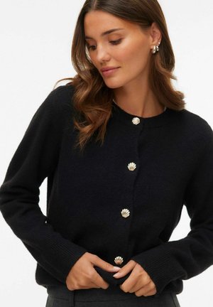 Frau trägt eine schwarze Strickjacke mit goldenen, muschelförmigen Knöpfen, richtet den Saum, lockiges, offen getragenes Haar und kleine silberne Ohrringe.