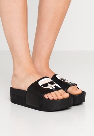 Feet wearing black platform slide sandals with cartoon faces of a person wearing sunglasses on the straps.