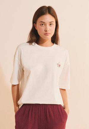 Giovane donna con capelli castani lisci che indossa una maglietta bianca con un piccolo ricamo floreale e pantaloni bordeaux strutturati, in piedi con le mani in tasca.