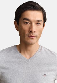 Asiatischer Mann mit kurzen dunklen Haaren, der ein hellgraues V-Neck-Shirt mit einem kleinen bestickten Logo auf der Brust trägt.