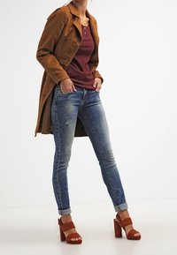 Kvinna i brun mockakappa, vinröd topp, uppvikta blå jeans och rostfärgade högklackade sandaler, står med händerna i fickorna.