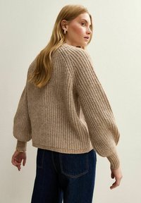 Pull en maille taupe à coupe décontractée, avec des manches à poignets côtelés et un motif texturé. Associez-le à un jean bleu foncé pour un contraste.