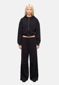 Zwarte cropped hoodie met een rits aan de voorkant en een trekkoord, gecombineerd met losse zwarte sweatpants. De stof lijkt zacht met een gladde textuur.