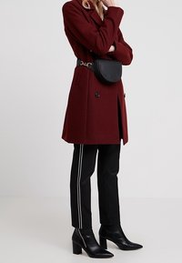 Bordová vlněná směsová bunda s dvojitým zapínáním, černá kabelka se zlatými akcenty, přiléhavé černé kalhoty s bílými bočními pruhy, černé boty na podpatku.