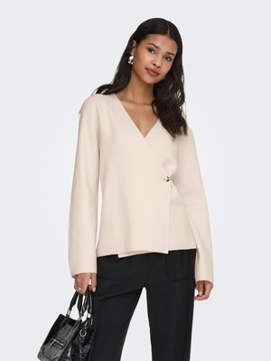 Kvinde iført beige omslagsstil langærmet bluse, sorte bukser, sølvøreringe og holder en sort håndtaske mod en ensfarvet baggrund.