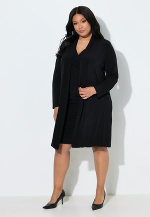 Vrouw met lang zwart haar, gekleed in een zwarte jurk tot op de knie, een zwarte lange cardigan en zwarte hoge hakken, staand tegen een witte achtergrond.