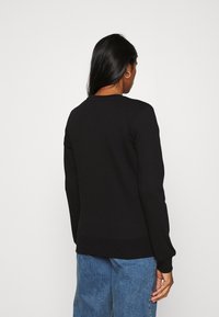 Mujer con cabello largo y negro, usando pendientes plateados en forma de aro, una camiseta negra de manga larga y jeans azules, de espaldas contra un fondo blanco liso.