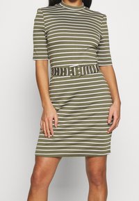 Kostium w kolorze oliwkowym w paski z białymi poziomymi paskami, krótki rękaw i dopasowany pasek z prostokątną klamrą. Dopasowany krój.