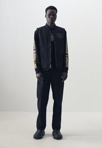 Veste noire zippée avec logo sur la poitrine et texte jaune sur les manches, associée à un t-shirt noir, un pantalon noir et des chaussures noires à semelles épaisses.