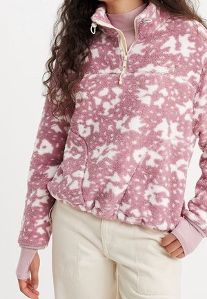 Femme portant un pull polaire à motifs rose et blanc avec une demi-fermeture éclair et un pantalon beige, sa main gauche dans la poche.