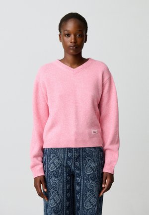 Mujer con un suéter rosa de cuello en V y pantalones azules con estampado, de pie frente a un fondo gris claro liso.