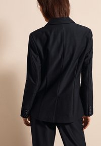 Chaqueta negra ajustada con una textura suave, solapas afiladas y puños abotonados. Presenta un diseño ceñido con detalles en la costura trasera.