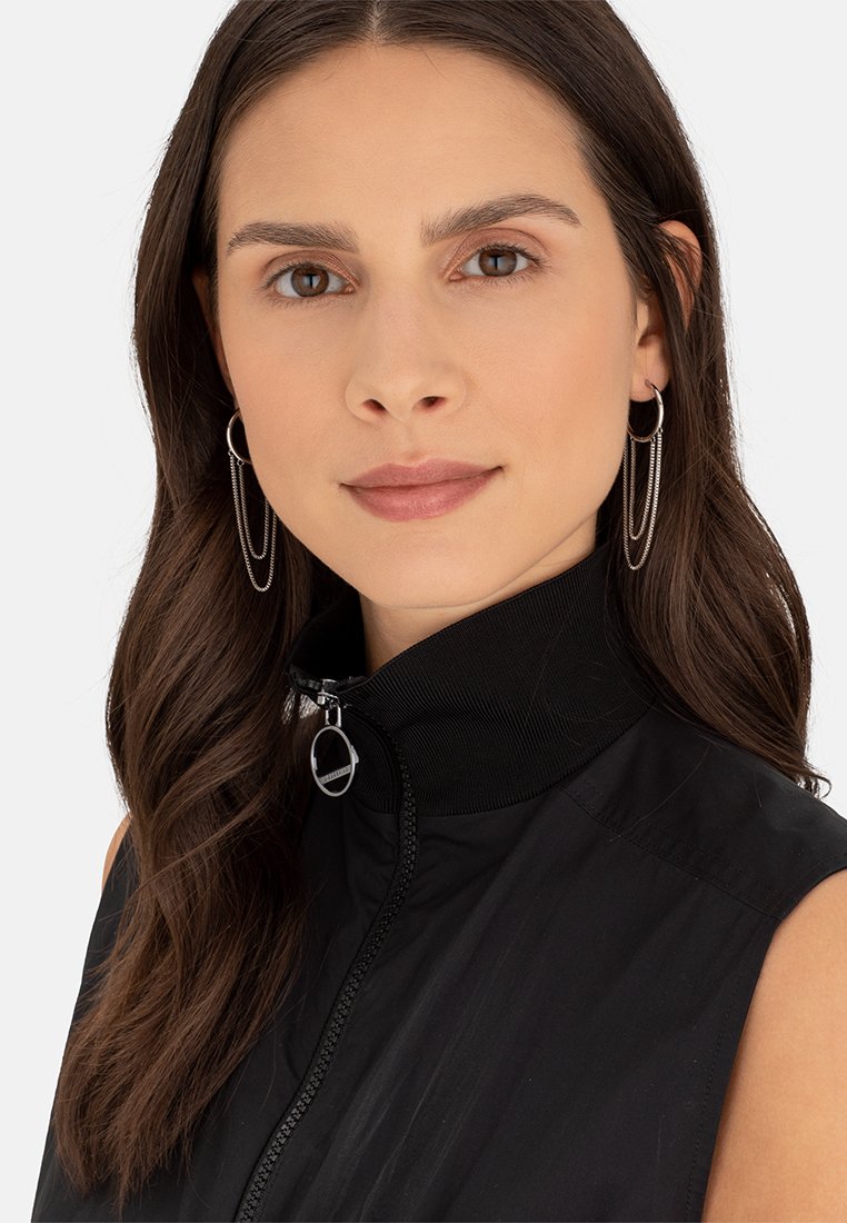 Zwart mouwloos jack met rits en hoge kraag, gemaakt van een textuurstof en voorzien van zilveren hardware, gedragen door een model met lang bruin haar.