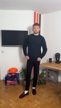 Σκούφο μπλε πουλόβερ με ριμπ μανσέτες, μαύρο εφαρμοστό παντελόνι και σκούρα καφέ παπούτσια. Το δωμάτιο διαθέτει πολύχρωμες πινελιές στους τοίχους και μοντέρνα διακοσμητικά στοιχεία.