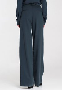 Szersze spodnie w ciemnoniebieskim materiale, wyróżniające się gładką fakturą, dwoma tylnymi kieszeniami i dopasowanym w talii krojem.