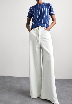 Osoba ubrana w niebiesko-białą koszulkę w paski z krótkim rękawem, włożoną w białe spodnie z wysokim stanem i szerokimi nogawkami, ze sznurkiem w pasie.