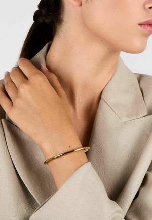 Braccialetto a bangle in oro indossato su un polso nudo, che riflette la luce. La persona è vestita con un blazer texturizzato di colore chiaro.