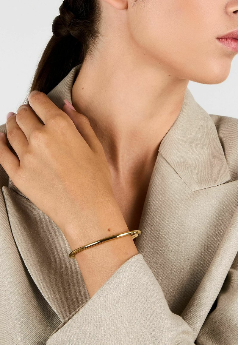 Braccialetto a bangle in oro indossato su un polso nudo, che riflette la luce. La persona è vestita con un blazer texturizzato di colore chiaro.