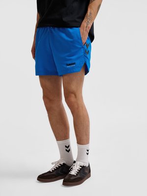 Mann trägt blaue Hummel-Sportshorts, weiße Socken mit schwarzen Chevron-Logos und schwarze Sneakers mit weißen Schnürsenkeln, steht mit einer Hand in der Tasche.
