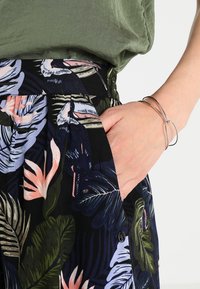 Gonna floreale con sfondo nero, decorata con grandi foglie verdi e fiori rosa. Dettaglio della tasca visibile, abbinata a un bracciale d'argento.