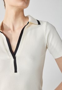 Ribbed short-sleeve top in cream with a black-trimmed V-neck collar; features textured fabric and a fitted silhouette.