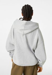 Persona con cabello trenzado que lleva una sudadera oversized gris claro y pantalones marrones, vista desde atrás sobre un fondo blanco.